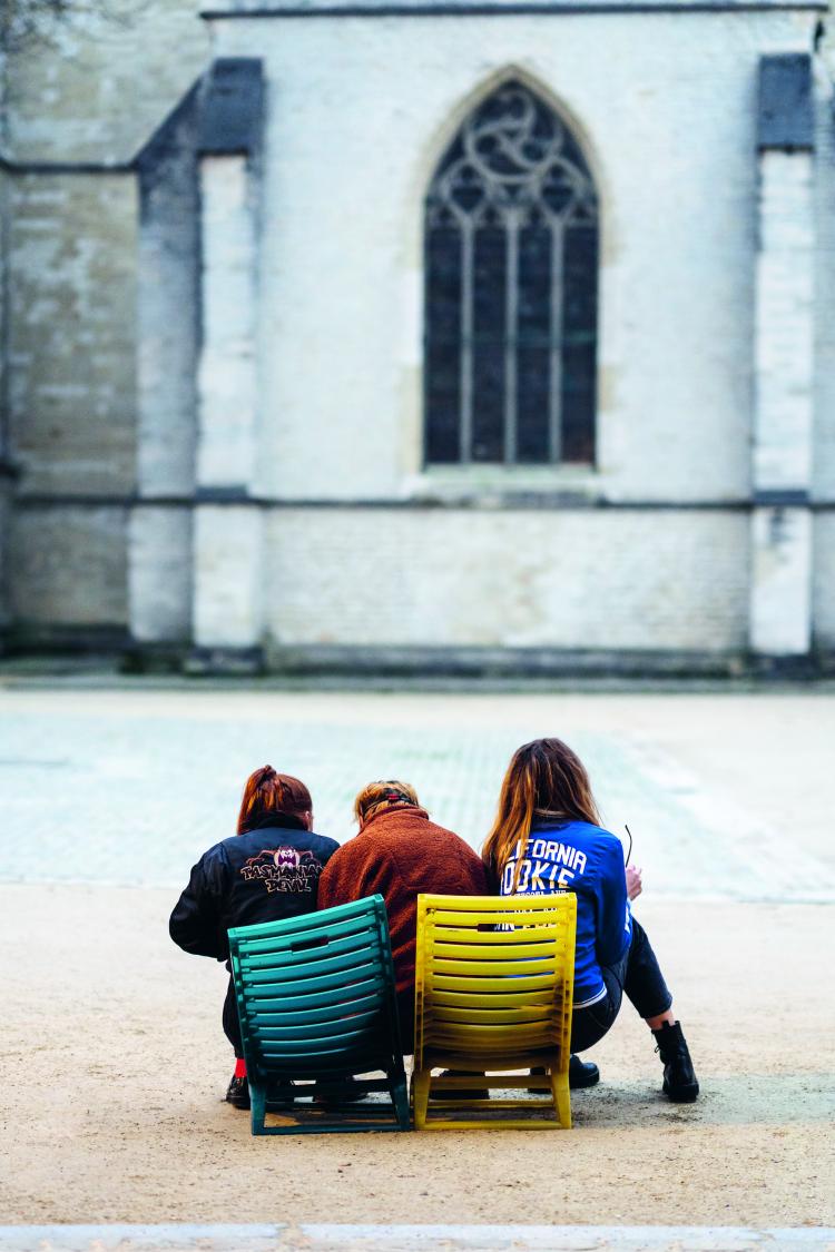 trois jeunes femmes assises de dos sur deux chaises de jardin colorées devant une église