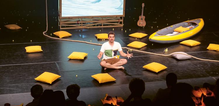 Photo d'un homme assis sur la scène et entouré de coussins jaune et d'un kayak